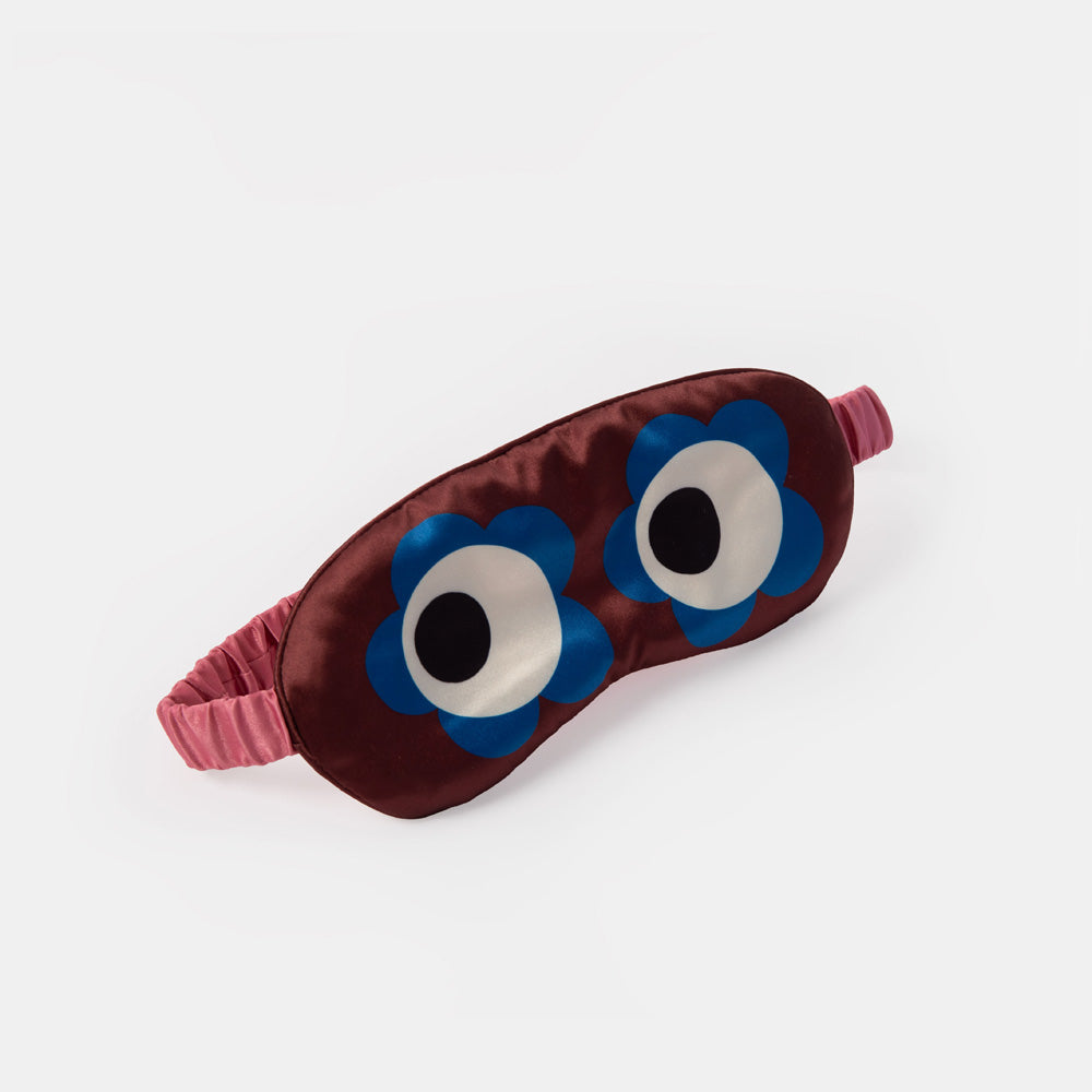 A dark red satin sleep mask featuring two large cartoon eyes with blue flower petals, white centers, and black pupils, complete with a pink elastic strap, set against a plain light gray background.