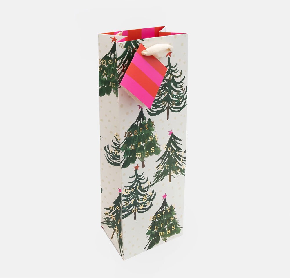 A tall Christmas-themed gift bag with a white background, decorated with green pine trees and gold 'MERRY CHRISTMAS' text, featuring a pink and red striped paper tag with a white ribbon handle.