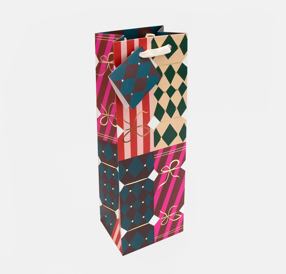 A tall, narrow gift bag with a geometric pattern of pink, teal, red, and beige Christmas crackers designs, featuring a white paper hanging loop at the top, shown on a light gray background.