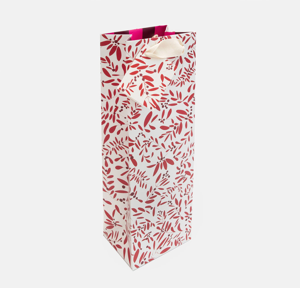 A tall white gift bag with an all-over red and dark gray botanical leaf pattern, featuring cream-colored paper handles and a bright pink ribbon at the top, shown on a light gray background.