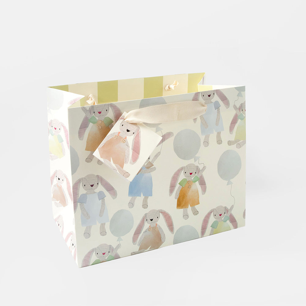 A large landscape gift bag with a pastel bunny print, featuring illustrated bunnies in soft colors on an off-white background, with a cream ribbon bow tied around the handle.