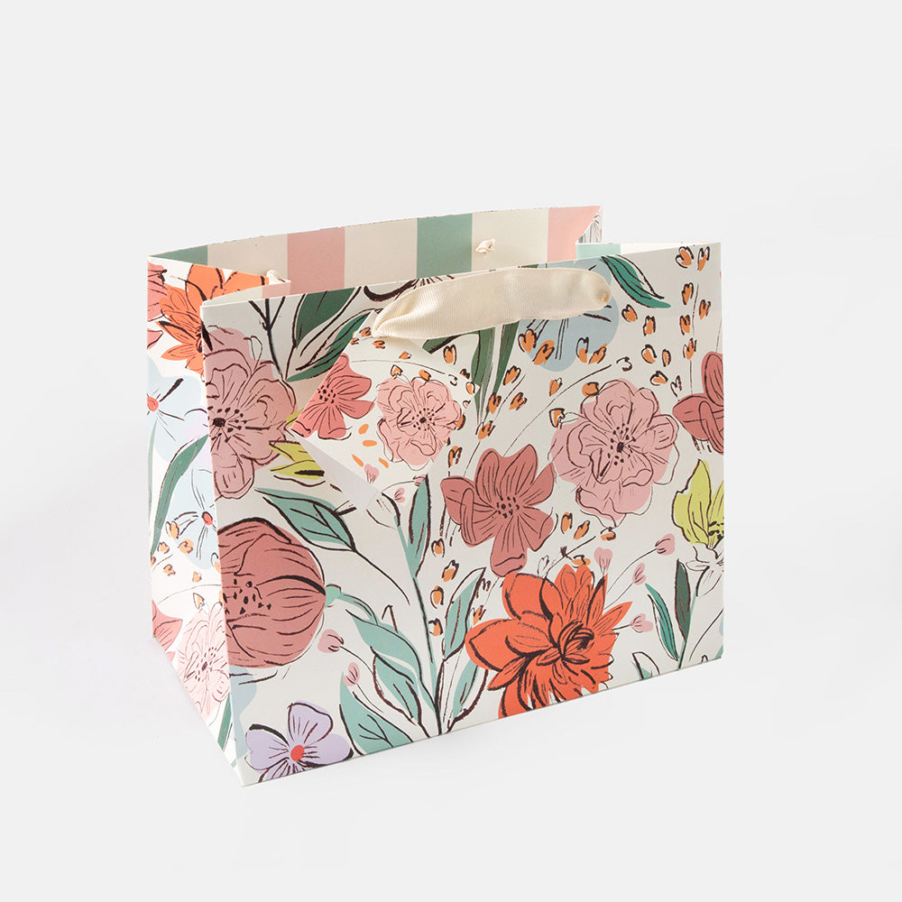 A white gift bag with a colorful floral pattern featuring pink, orange, and green flowers, with a striped pattern on the top edge and a cream-colored paper handle.