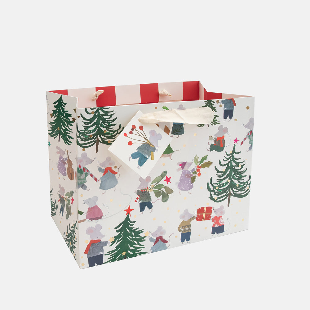 A white gift bag with a festive Christmas print featuring mice in holiday outfits, evergreen trees, and small gift boxes, complete with red striped inner lining and cream paper handles.