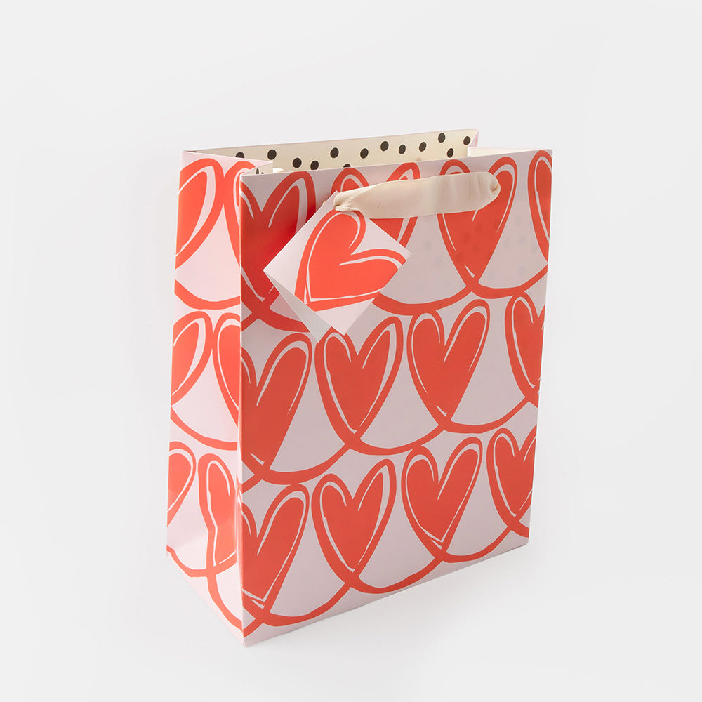 A gift bag with a repeating pattern of coral-red hearts on a white background, featuring cream paper handles with black polka dots.