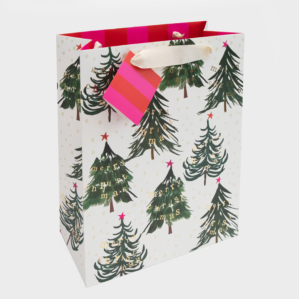 A white gift bag with a festive Christmas tree pattern, featuring green pine trees with gold 'Merry Christmas' lettering and pink accents on the handles and inner lining, set against a light gray background.