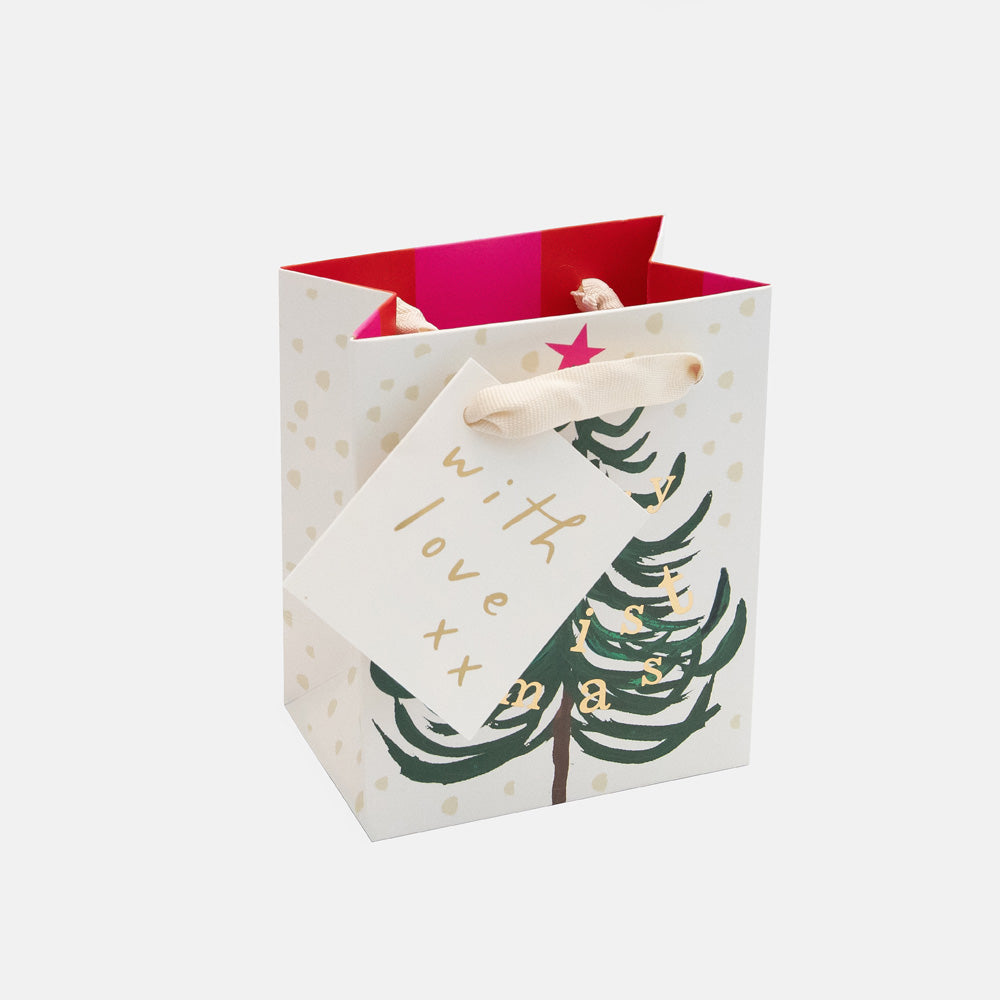 A white gift bag with gold polka dots, a green Christmas tree illustration, and a hanging tag that reads 'with love xx' in gold script. The bag has a bright pink interior and is shown on a light gray background.