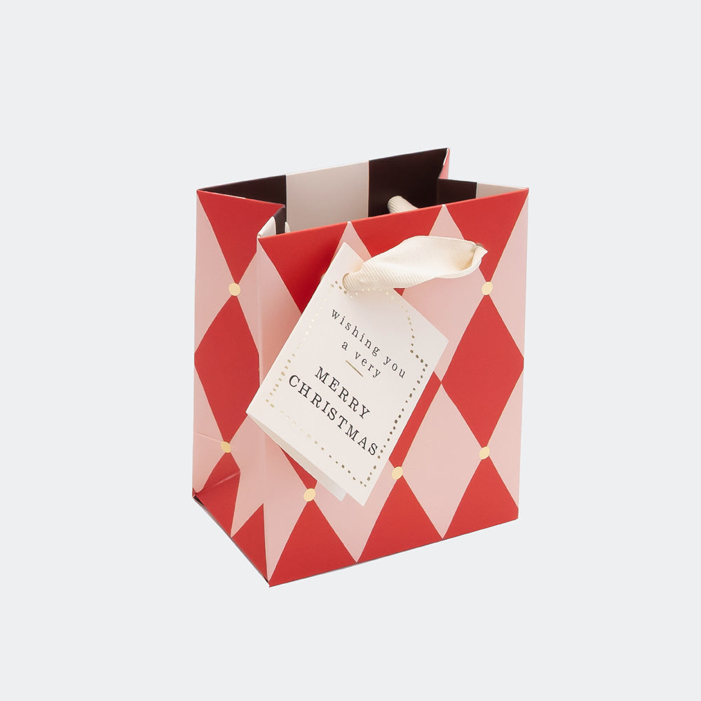 A red and light pink geometric patterned Christmas gift bag with a white ribbon and a hanging tag that reads 'MERRY CHRISTMAS'.