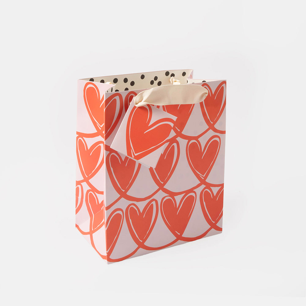 A small gift bag with a repeating pattern of coral-red hearts on a white background, featuring cream handles with black polka dots.