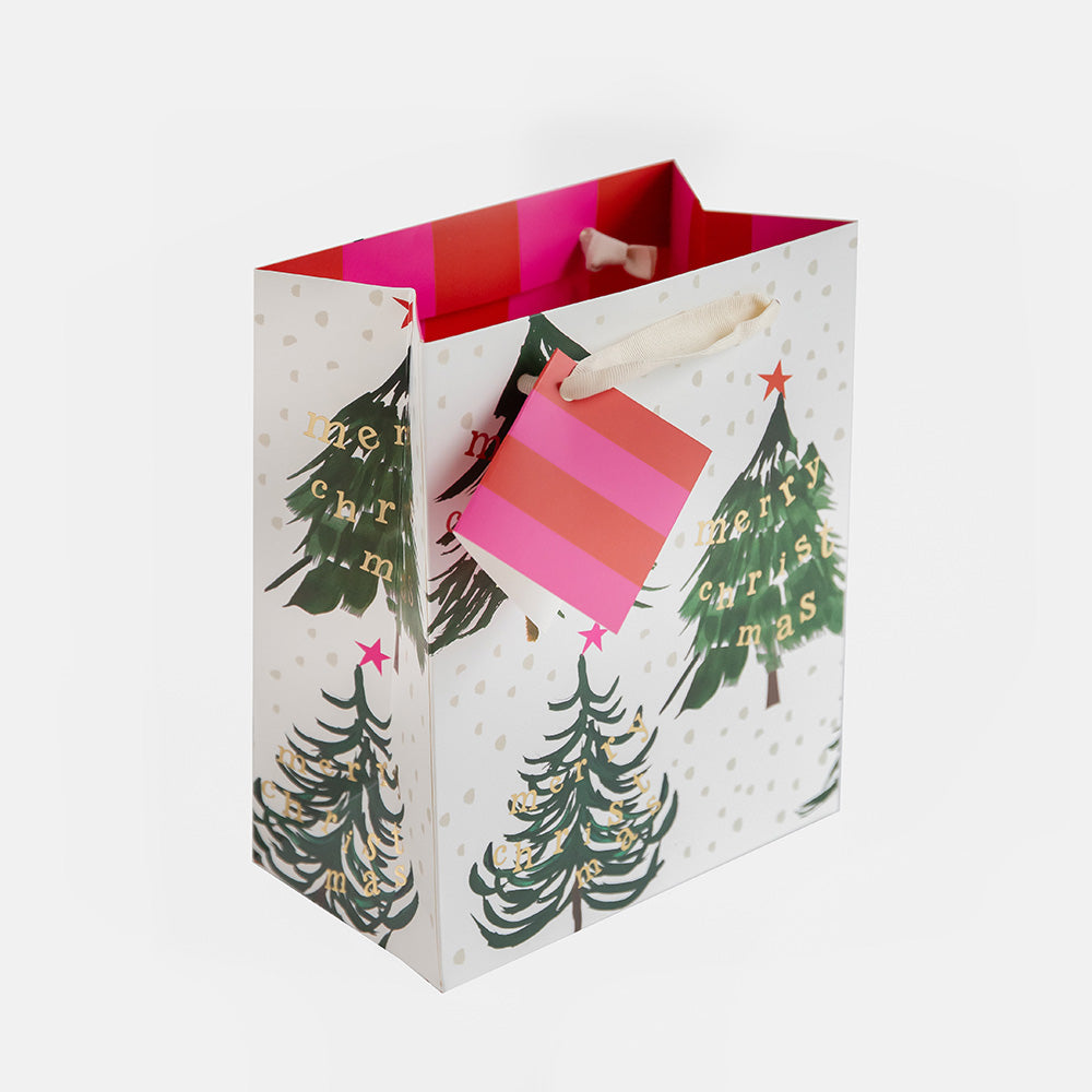 A white Christmas-themed paper gift bag with green tree illustrations, gold lettering, and pink striped handles and tag, shown on a light gray background.
