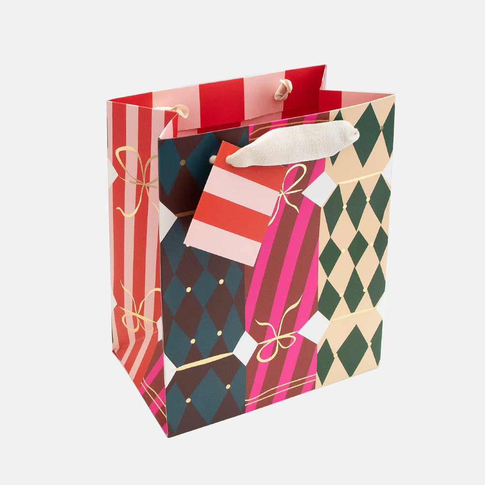 A festive paper gift bag with a multicolor christmas cracker design featuring red and white stripes, pink and green harlequin diamonds, and gold bow accents, complete with white rope handles and a matching red and white gift tag.