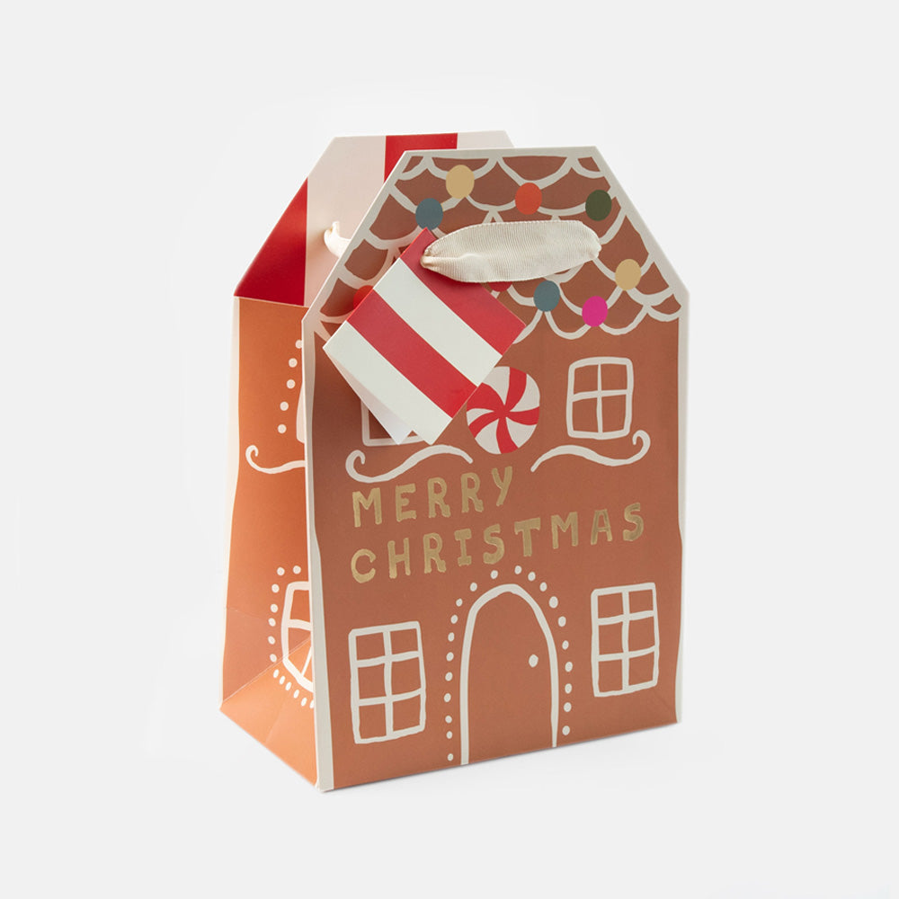 A gingerbread house shaped Christmas gift bag with 'MERRY CHRISTMAS' printed on the front, featuring a red and white striped candy cane and colorful holiday lights, set against a plain white background.
