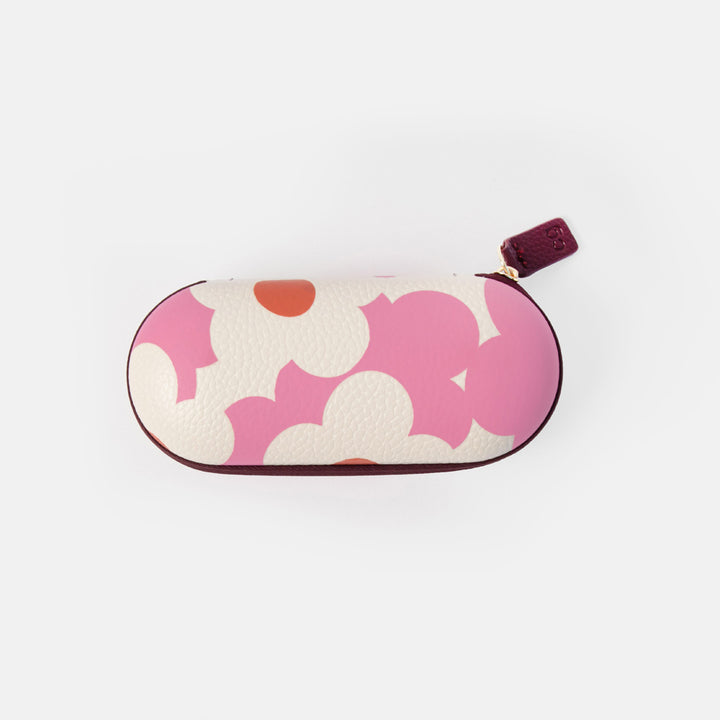 An oval-shaped eyeglasses case featuring a pink and white floral pattern with a small orange accent, finished with a dark burgundy leather zipper pull tab.