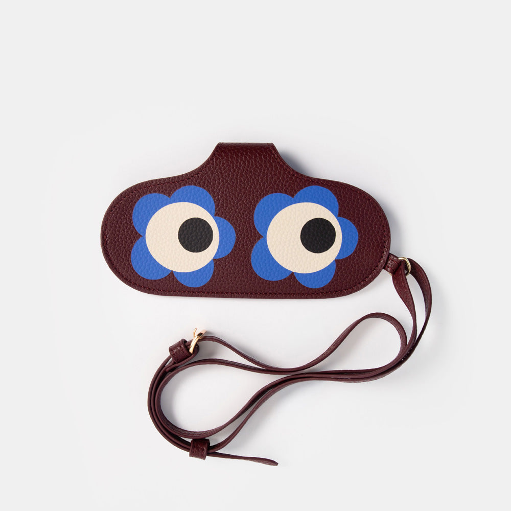 A dark brown pebbled leather wristlet glasses case with a strap, featuring a playful cartoon face made of two large blue flower petals and cream-colored eyes with black pupils.