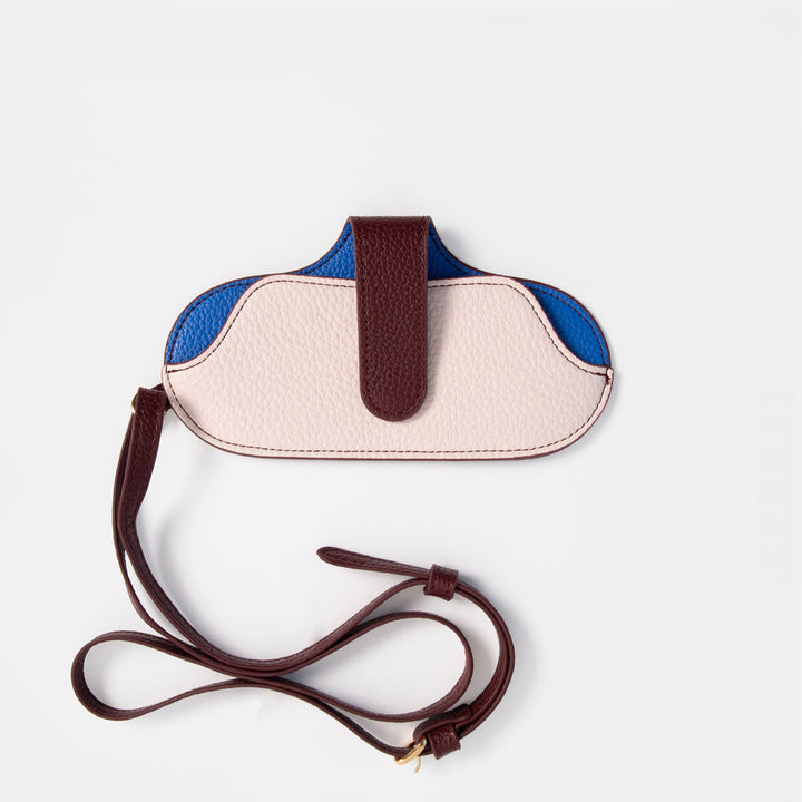 A color-blocked glasses case with a curved shape, featuring a light pink pebbled leather front, blue leather side panels, and a dark brown strap and central tab, displayed on a light gray background.