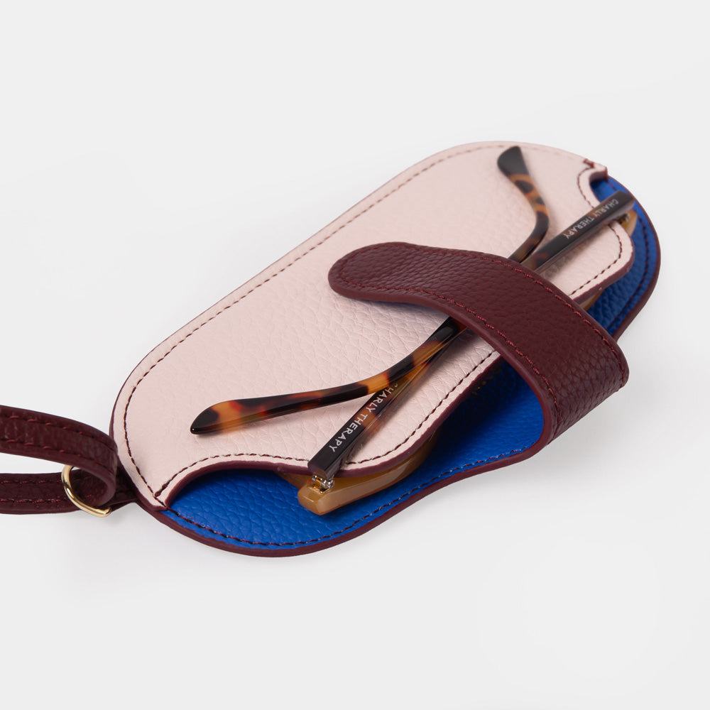 A pair of tortoiseshell eyeglasses resting on a light pink and blue flip-down case with a burgundy strap, photographed on a clean white background.