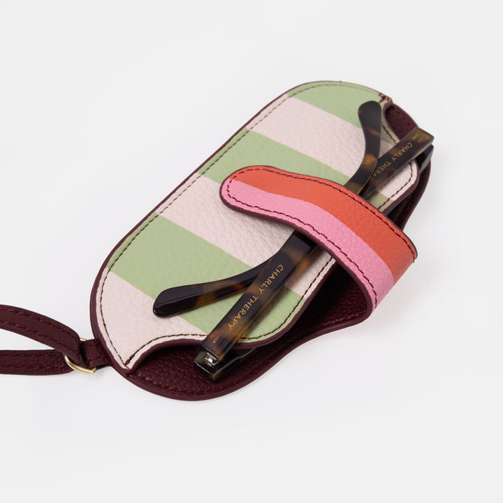 A pair of tortoiseshell sunglasses resting on a striped leather sunglasses case with a matching wrist strap. The case features horizontal stripes of sage green, light pink, and white, with dark burgundy leather trim and a bright pink and coral strap.
