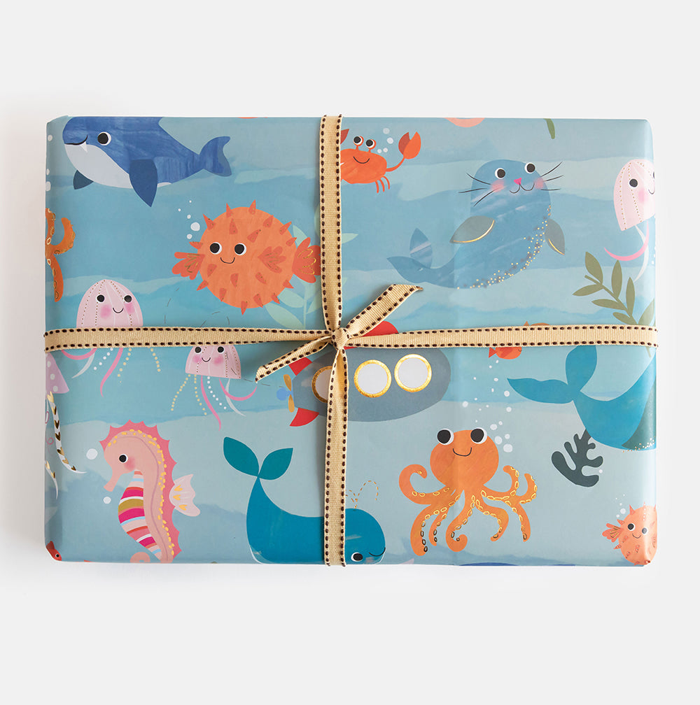 A gift wrapped in light blue wrapping paper with a colourful cartoon ocean animal print, tied with a beige ribbon and bow, on a white background.