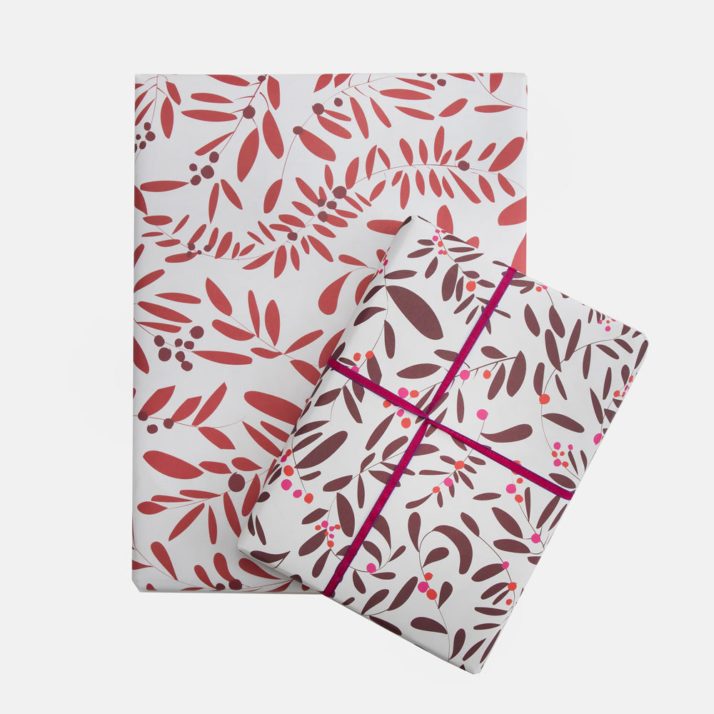 Two rectangular pieces of wrapping paper with a botanical leaf pattern, shown on a light gray background. One flat sheet and one wrapped rectangular item, both featuring a white base with red and brown leaves and small berry motifs, tied with a magenta ribbon.