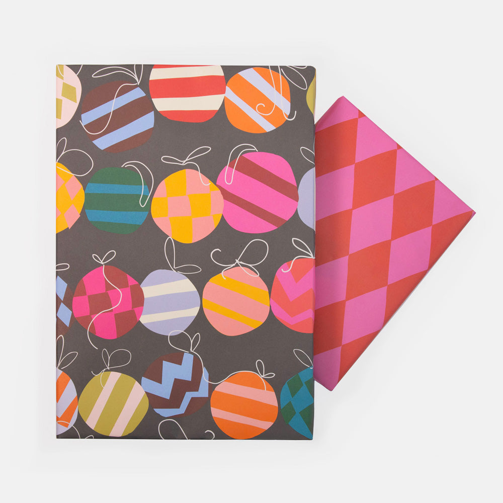 Christmas wrapping paper sheets, one with a dark grey design featuring a colourful pattern of striped and checked Christmas ornaments, one with pink and red diamond-pattern on a clean white background.