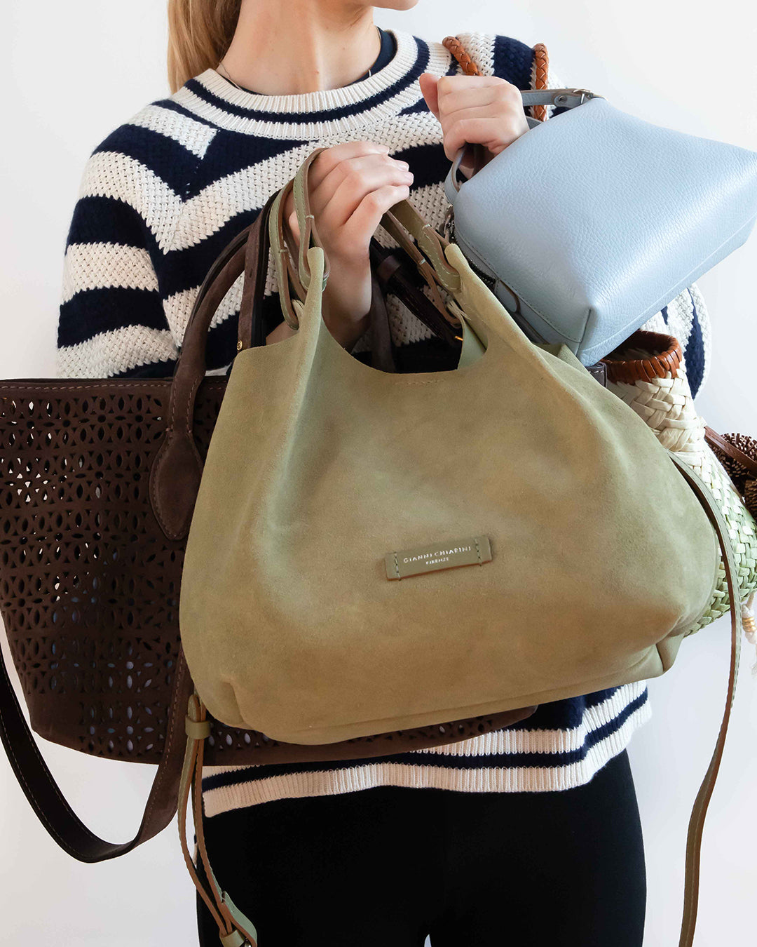 A person wearing a navy and cream striped sweater holds a large olive green suede hobo bag in front of a light blue handbag and a brown woven tote bag. The brand name 'MARC JACOBS' is visible on a small metal plaque on the front of the green bag.