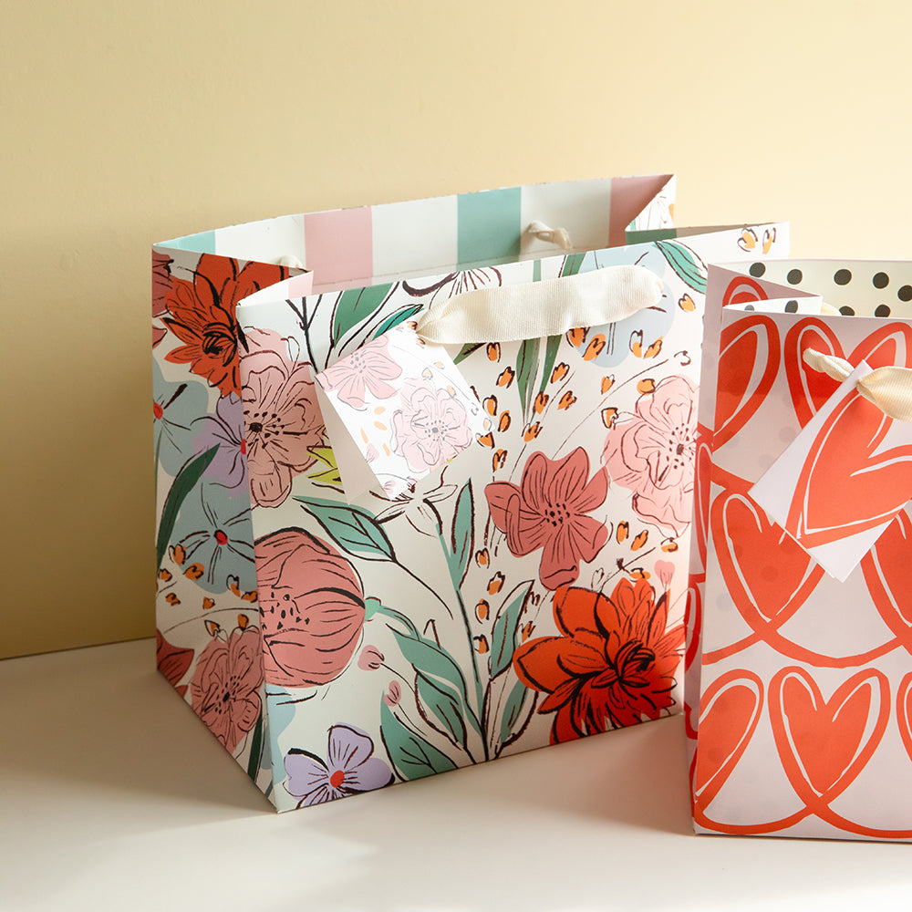 Two decorative gift bags with white backgrounds and colorful floral and heart patterns, displayed on a light surface against a warm beige wall.
