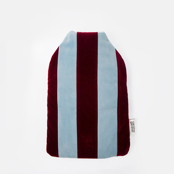 A plush velvet hot water bottle with vertical stripes in burgundy and light blue, featuring a small white sewn-in tag on the right side.