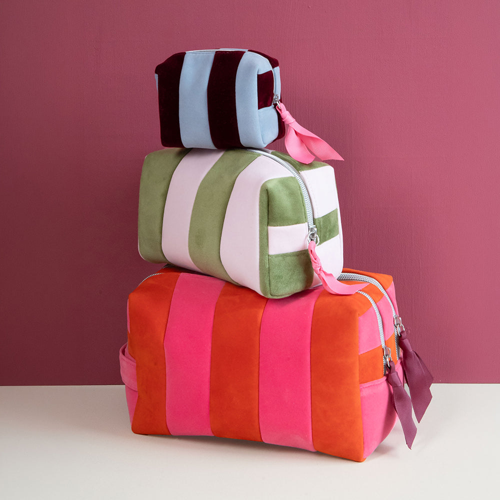 Three stacked striped fabric cosmetic bags in pink, orange, green, white, and navy, set against a solid dark pink background.