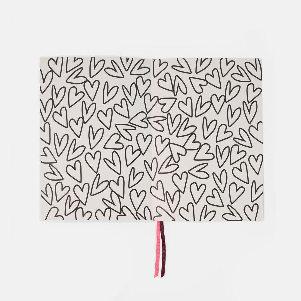 A rectangular white notebook or journal with an all-over black hand-drawn heart pattern, featuring a thin pink and dark red ribbon bookmark hanging from the bottom center, set against a plain light gray background.