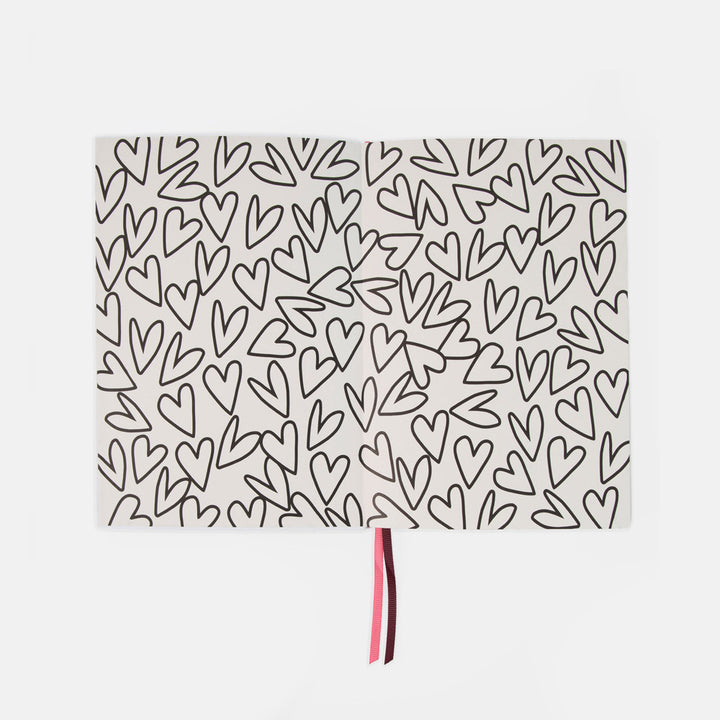 A rectangular white notebook or journal with an all-over black hand-drawn heart pattern, featuring a thin pink and dark red ribbon bookmark hanging from the bottom center, set against a plain light gray background.