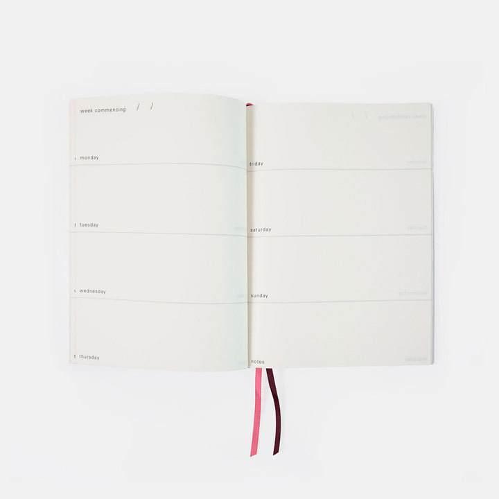 An open weekly planner with blank white pages, featuring two ribbon bookmarks in pink and dark red, shot against a solid white background.