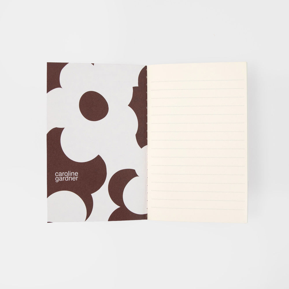 An open Caroline Gardiner lined notebook showing a white cover with a brown abstract floral pattern on the left and cream-colored lined pages on the right, set against a light gray background.