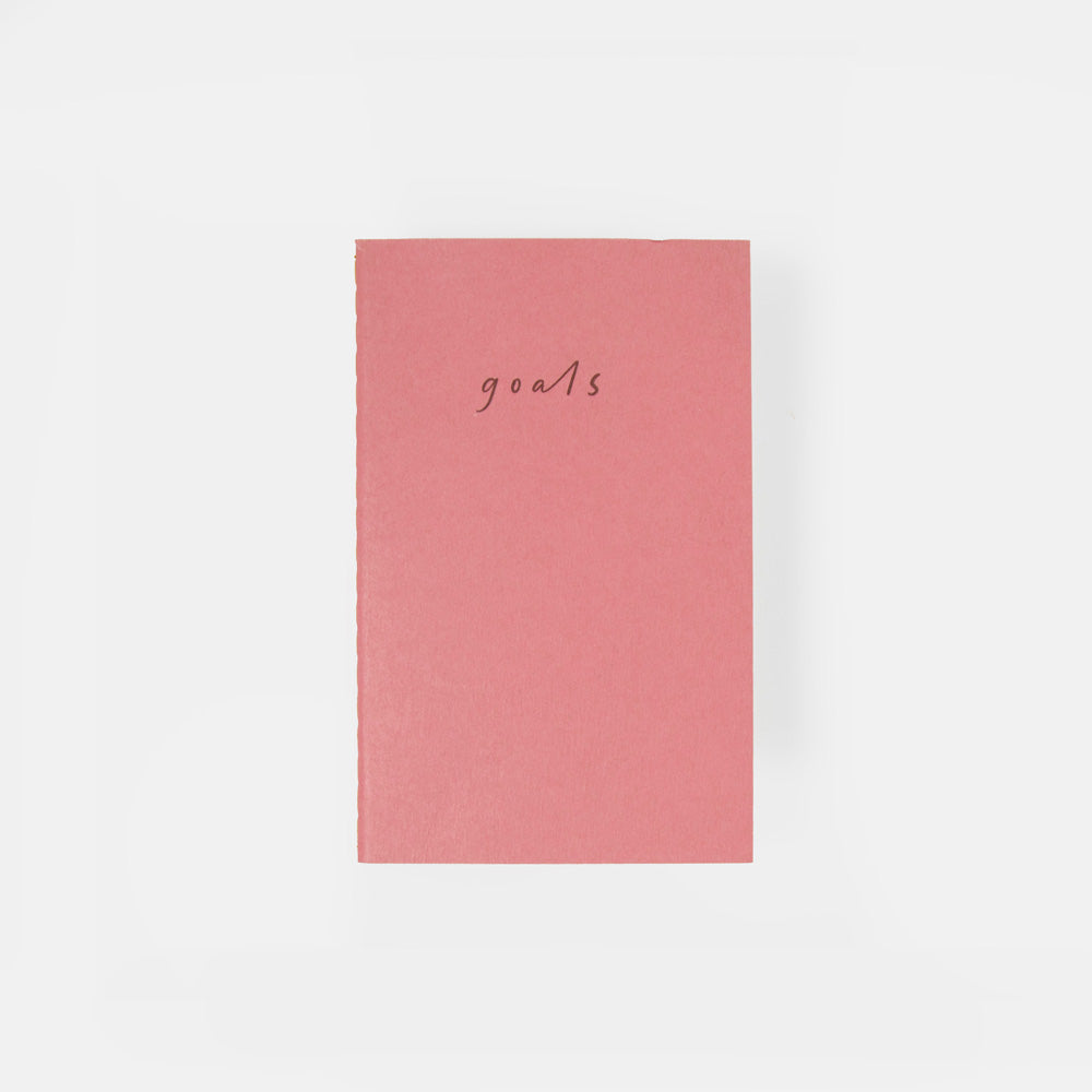 A pink hardcover notebook with the word 'goals' written in a small cursive font on the front, centered against a plain white background.