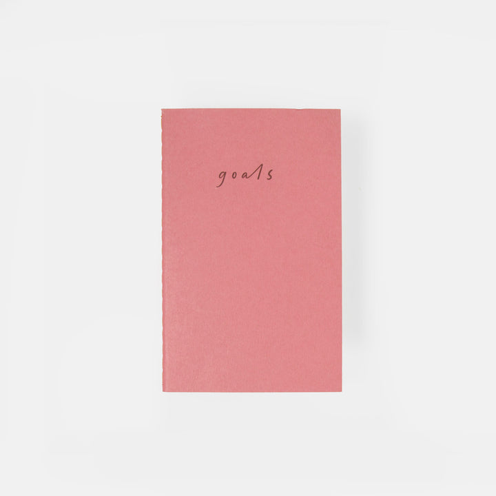 A pink hardcover notebook with the word 'goals' written in a small cursive font on the front, centered against a plain white background.