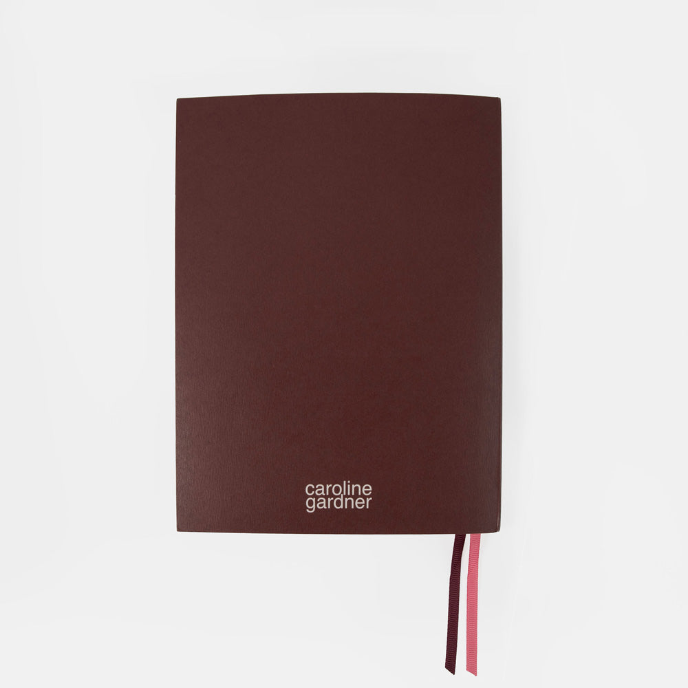 A dark brown hardcover notebook with the brand name 'caroline gardiner' printed in white near the bottom left, featuring a pink and dark brown ribbon bookmark on a light gray background.