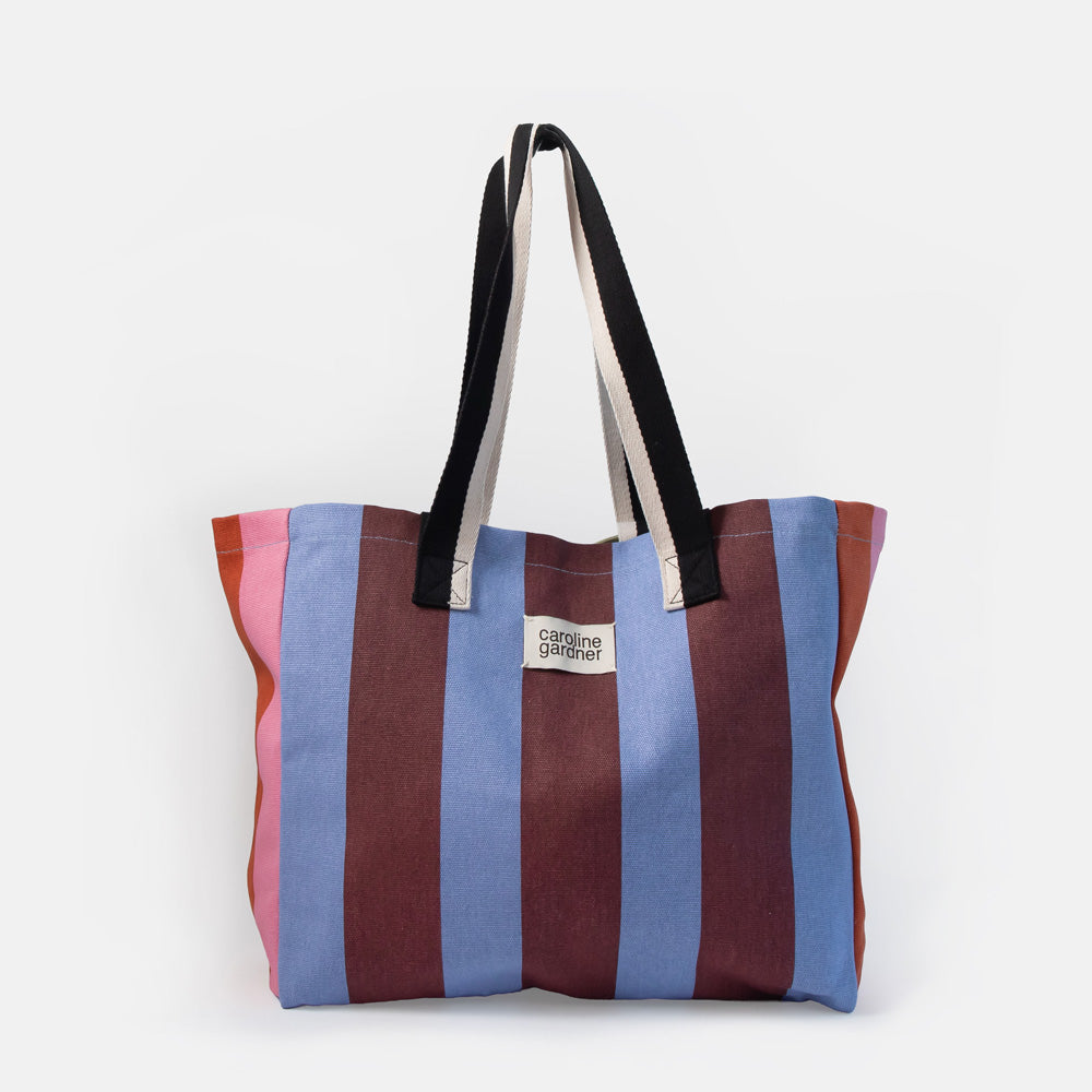 A striped canvas tote bag with vertical stripes in pink, blue, and burgundy, featuring black and white shoulder straps and a small white brand label that reads 'caroline gardner' on a light gray background.