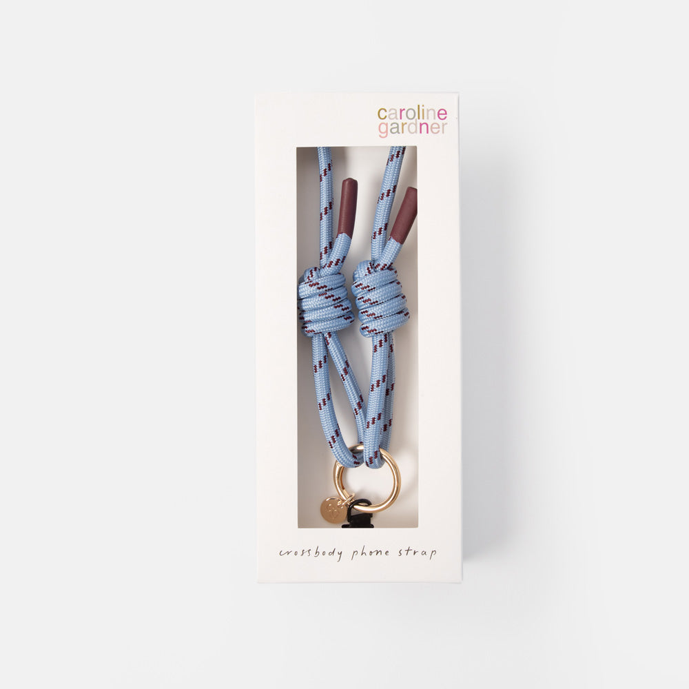 Caroline Gardner phone strap in its original white window box packaging, featuring a light blue paracord knot with purple and red stripes, a gold ring, and a small heart-shaped charm.