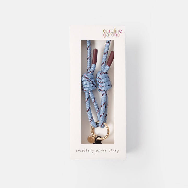 Caroline Gardner phone strap in its original white window box packaging, featuring a light blue paracord knot with purple and red stripes, a gold ring, and a small heart-shaped charm.