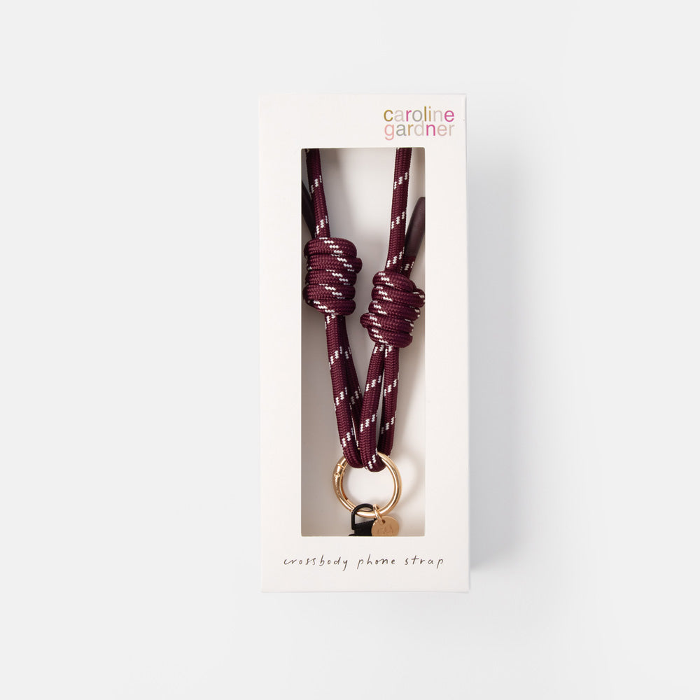 A Caroline Gardner phone strap in a deep burgundy color with white speckles, featuring knotted details and a gold-tone ring, shown inside its white retail packaging box against a light gray background.
