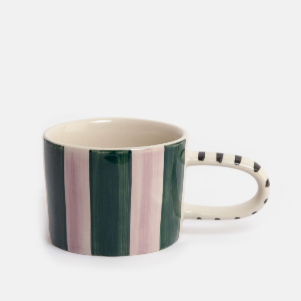 A ceramic mug with vertical stripes in dark green, light pink, and cream, featuring a cream handle with black polka dots, set against a plain light gray background.