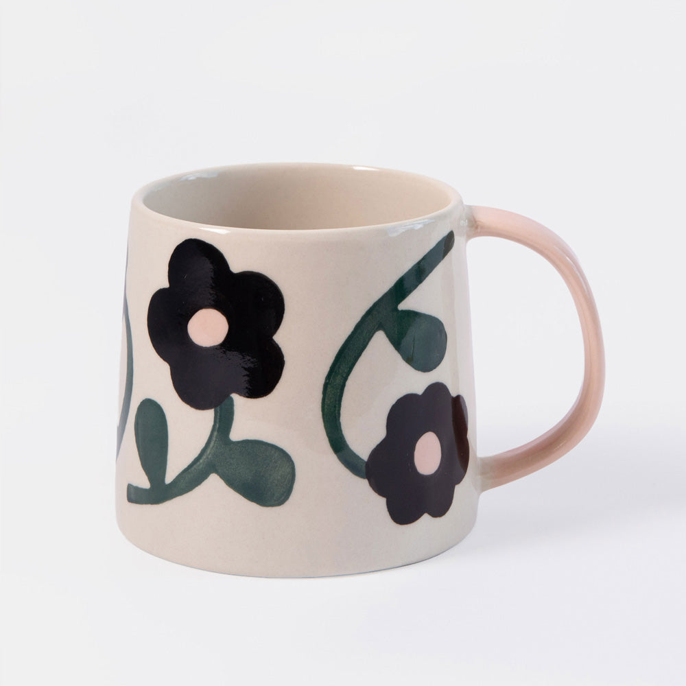 A ceramic mug with a warm cream-colored body, decorated with hand-painted black flowers and dark green leaves, featuring a contrasting light pink handle.