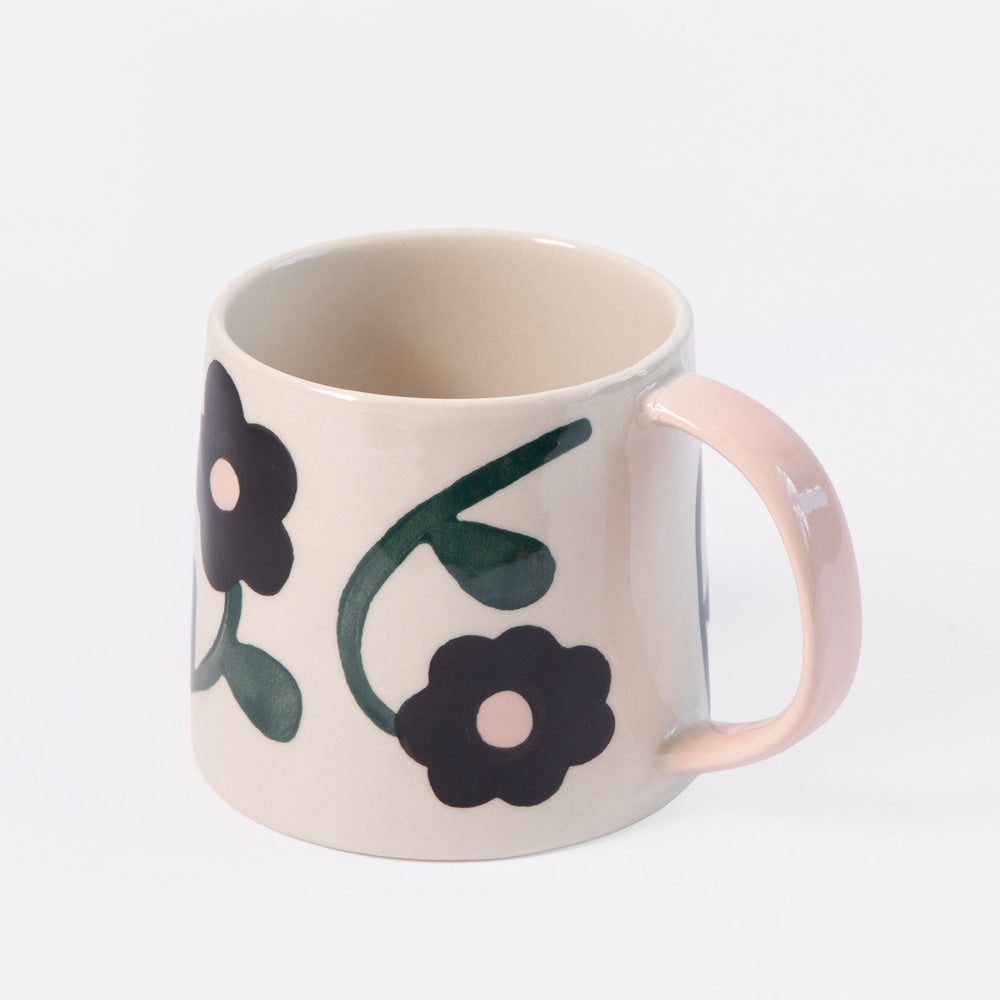 A ceramic mug with a warm cream-colored exterior, decorated with two black flowers and green leaves, featuring a contrasting light pink handle.