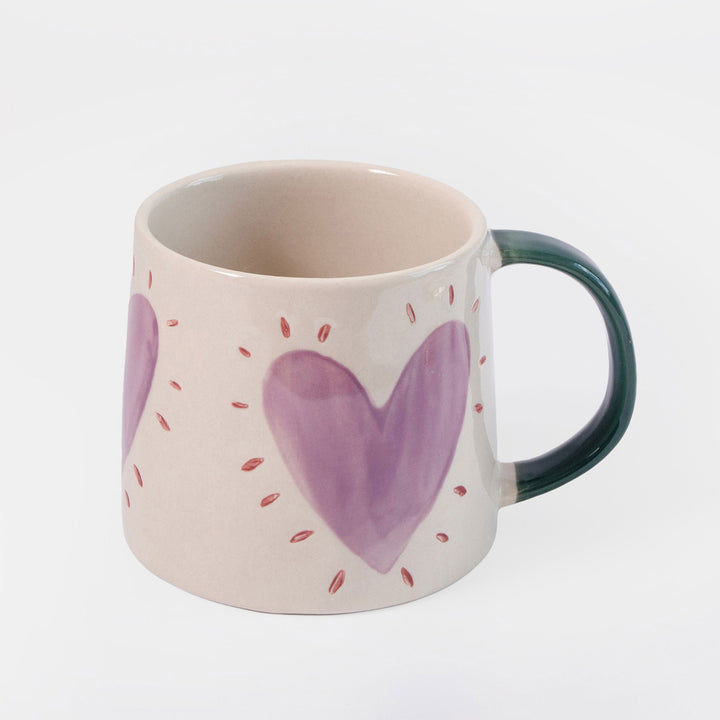 A ceramic mug with a light pinkish-beige exterior decorated with purple hearts and small red dotted lines, featuring a dark green handle.