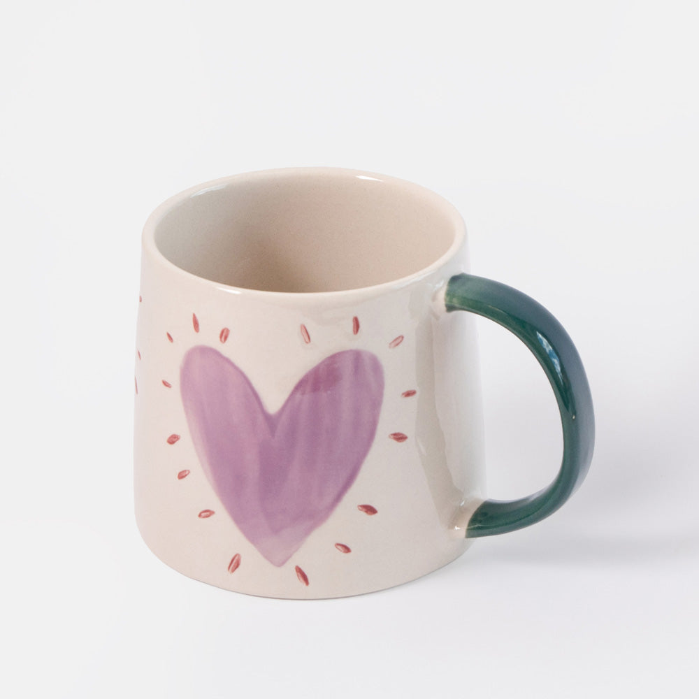 A ceramic mug with a cream-colored body, a hand-painted purple heart with small red dots, and a dark green handle, shown on a light gray background.