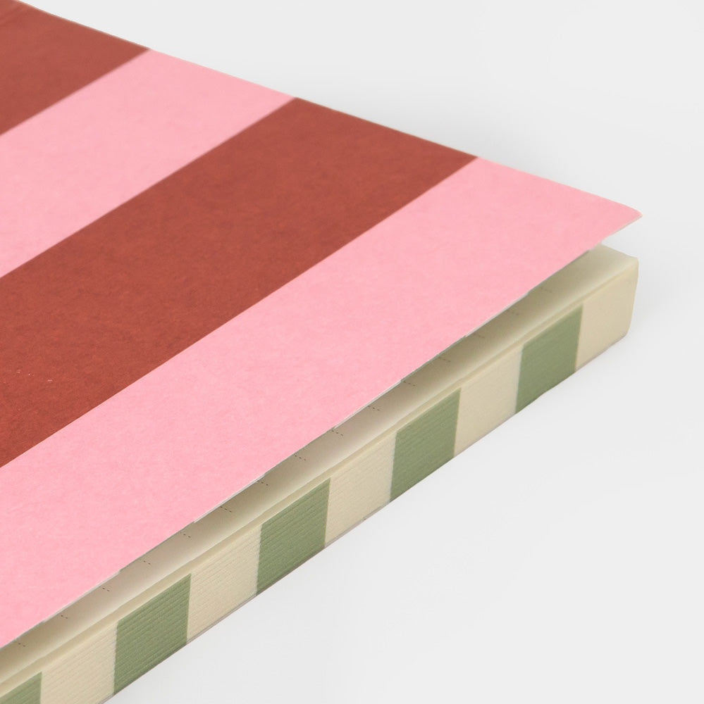 A close-up of a notebook or journal showing a striped cover with alternating pink and rust-red bands, a cream page block, and sage green side stripes, photographed on a light gray background.