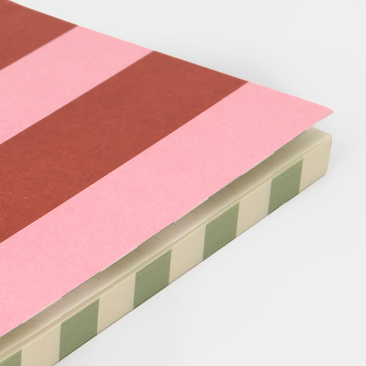 A close-up of a notebook or journal showing a striped cover with alternating pink and rust-red bands, a cream page block, and sage green side stripes, photographed on a light gray background.