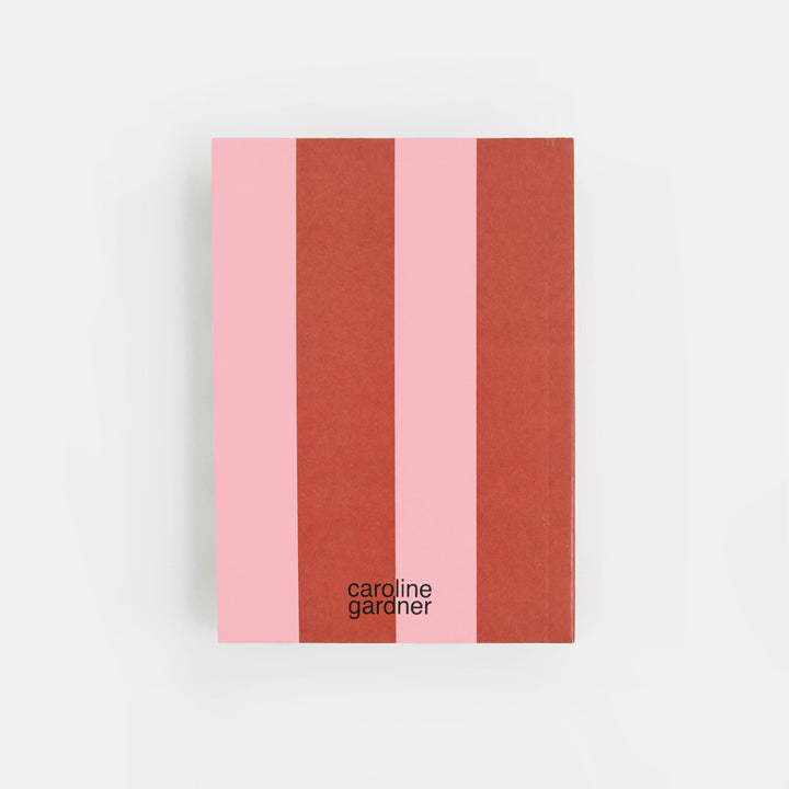 A notebook with a minimalist cover design featuring four vertical stripes in light pink, terracotta, and rust red, with the text 'caroline gardner' printed in black near the bottom, set against a clean white background.