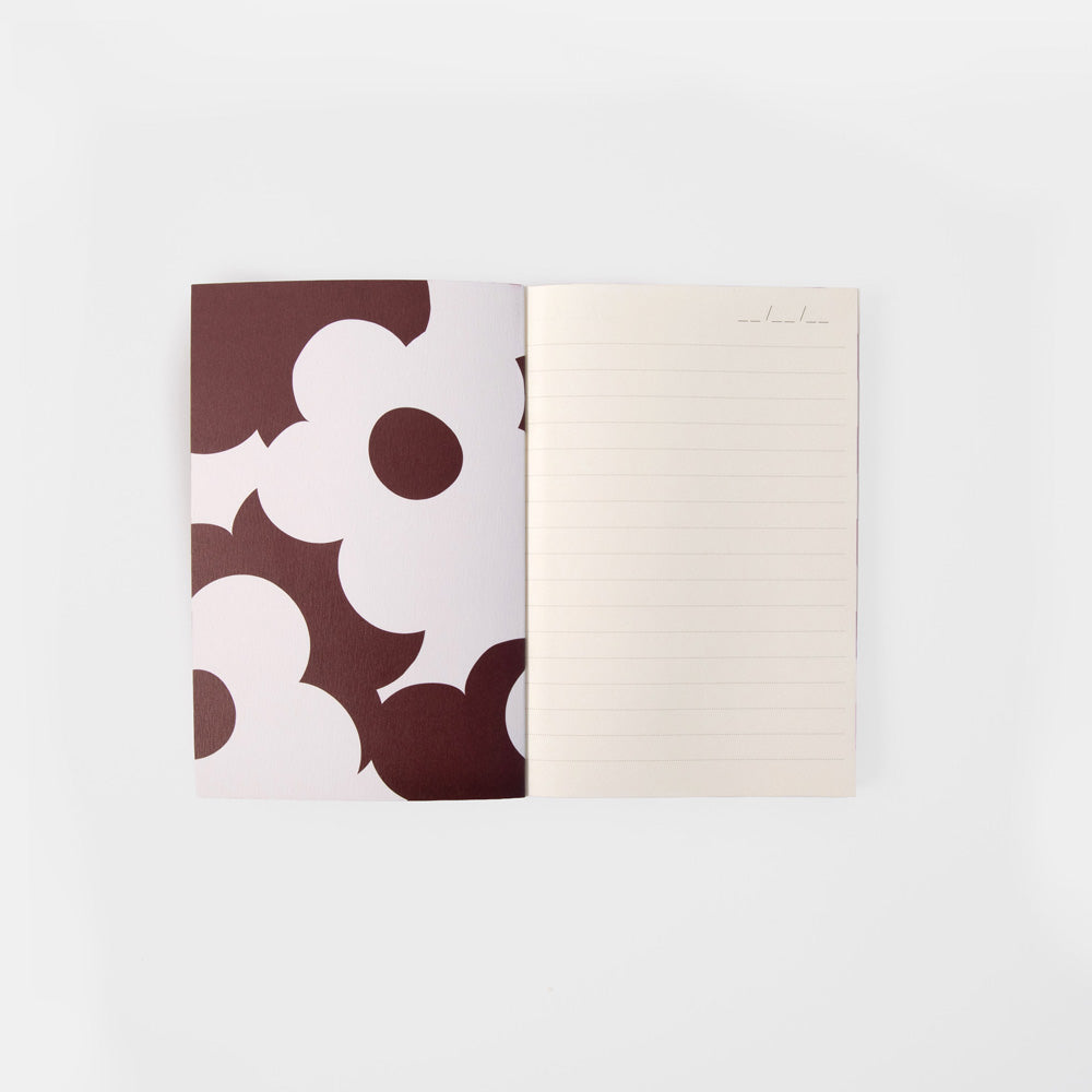 An open lined notebook with a cream page on the right and a cover featuring a bold white and dark brown floral pattern on the left, photographed on a clean white background.