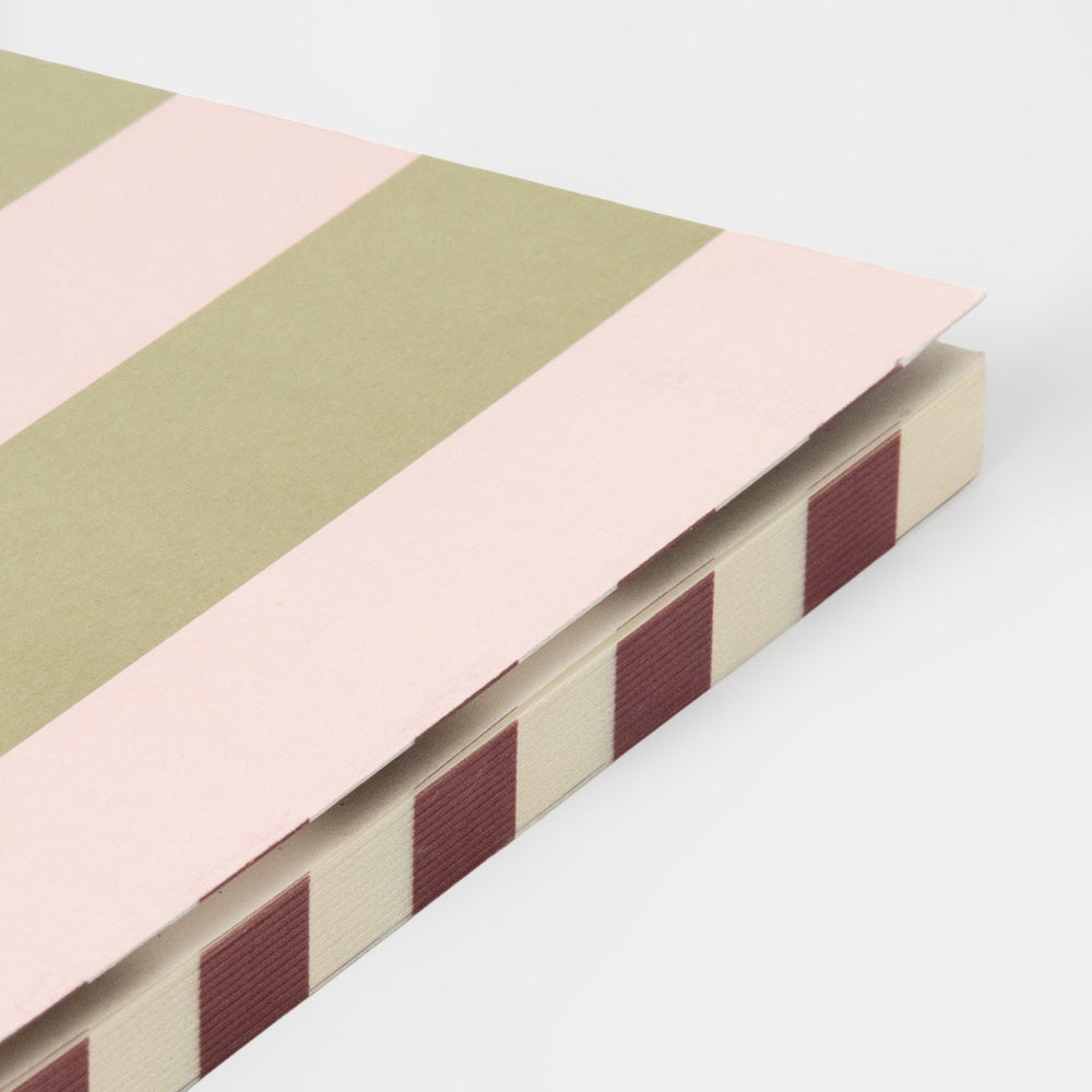 A close-up of a notebook or journal corner showing a striped cover pattern with dusty pink, sage green, cream, and dark red bands.