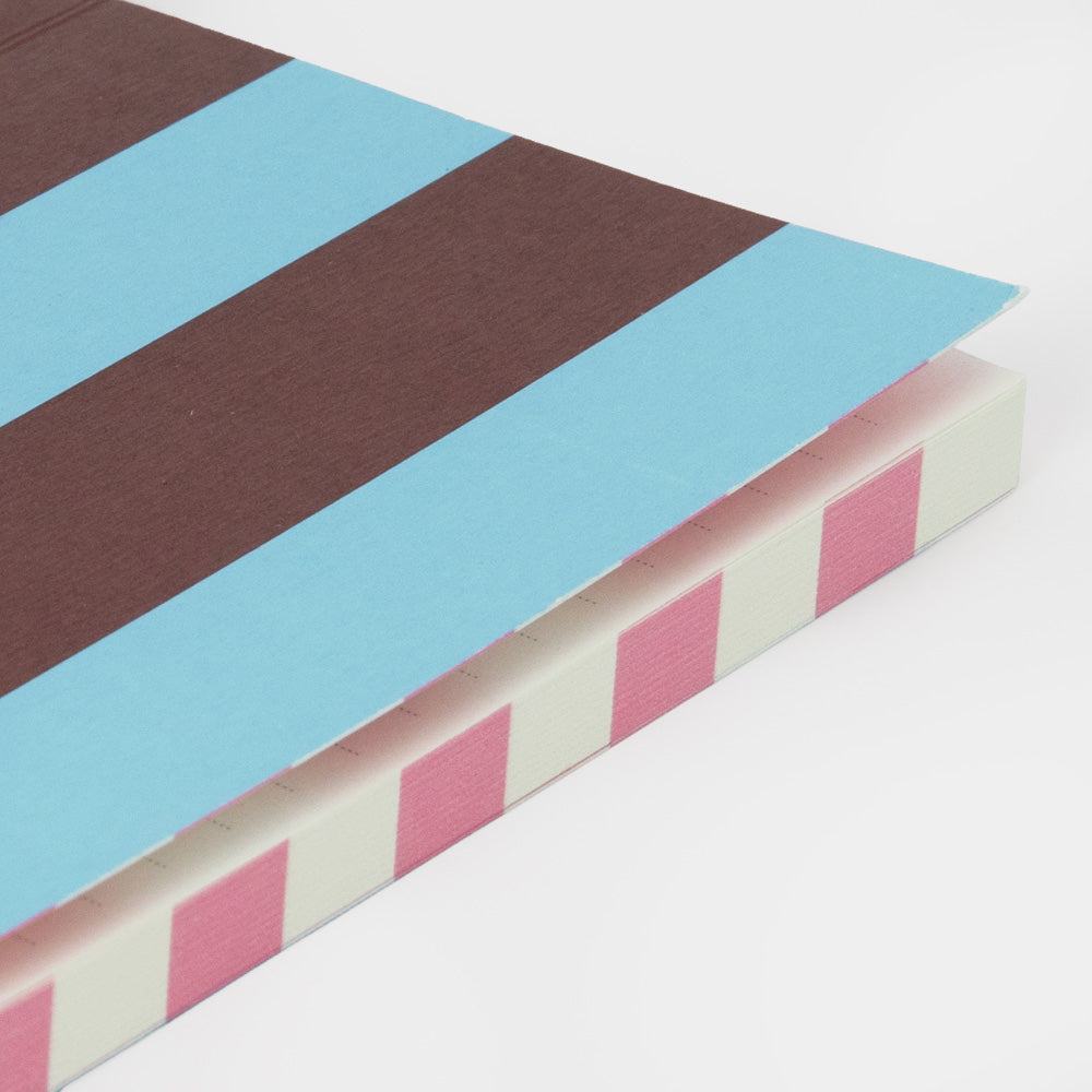 A close-up of a notebook or journal corner showing a striped cover design with wide light blue and dark brown bands on top, and alternating pink and off-white stripes along the side edge, photographed on a clean white background.
