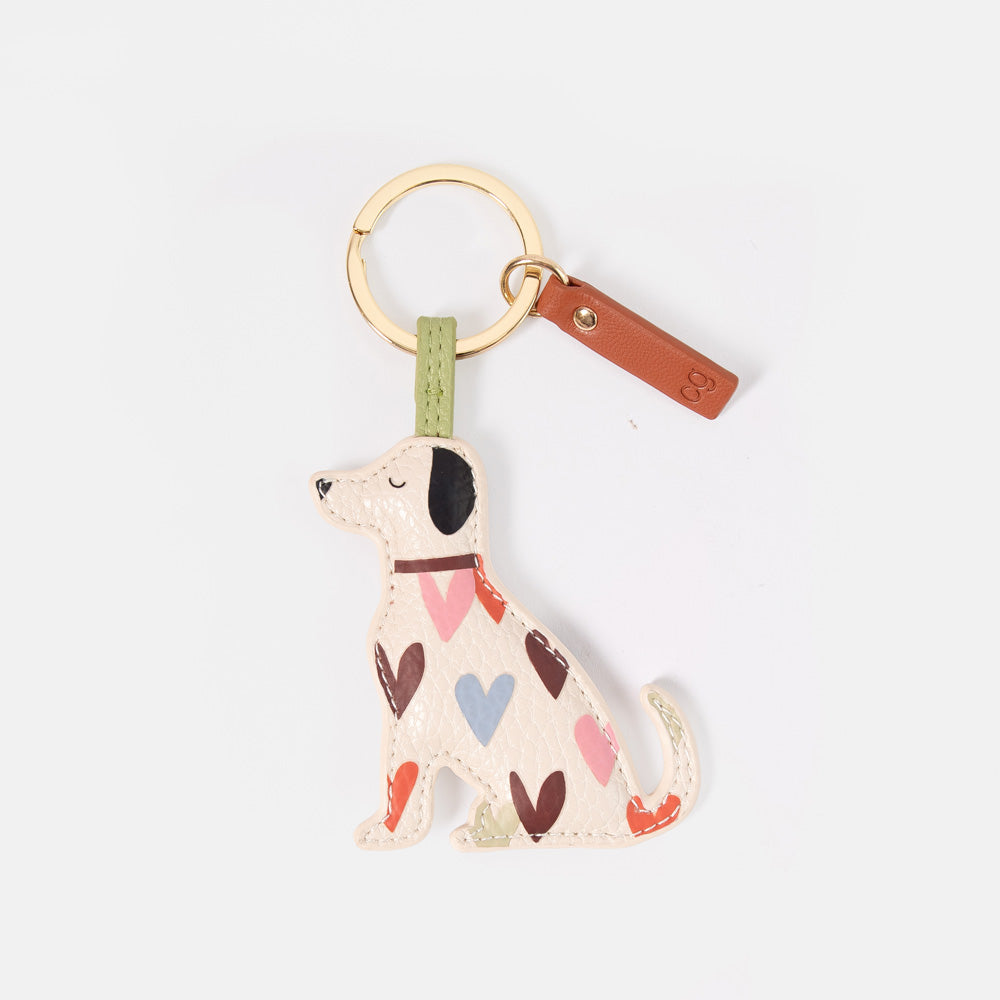 A dog-shaped keyring or bag charm, featuring a cream-colored body with a pattern of pink, red, brown, and blue hearts, attached to a gold-tone split ring and a brown leather tag.